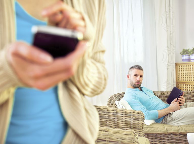 אִשָׁה texting someone on cell phone, curious husband in the background sitting in a chair